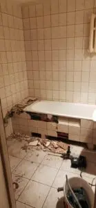 Łazienka w remoncie z częściowo rozebraną ścianą wyłożoną kafelkami, odsłaniającą leżące pod nią konstrukcje i gruz rozrzucony na podłodze.