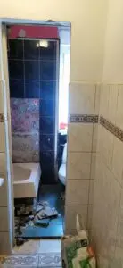 Łazienka w remoncie z gruzem na podłodze, częściowo usuniętymi płytkami na ścianie i odbiciem toalety w lustrze.