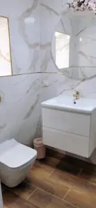 Nowoczesne wnętrze łazienki z marmurowymi płytkami, elegancką białą szafką podumywalkową, okrągłym lustrem i podłogą z efektem drewna.