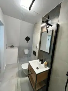 Elegancka i nowoczesna łazienka wyposażona w szklaną kabinę prysznicową, toaletę podwieszaną oraz toaletkę z prostokątną umywalką i dużym lustrem. paleta kolorów jest neutralna z mieszanymi teksturami na ścianach.