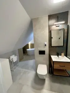 Nowoczesne i eleganckie wnętrze łazienki w neutralnych kolorach, z toaletą montowaną na ścianie, drewnianą toaletką z umywalką, szafką z lustrem, przeszkloną strefą prysznicową i wolnostojącą wanną pod skośnym sufitem.