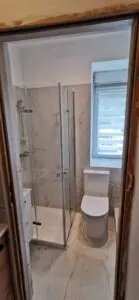 Nowoczesna łazienka z marmurowymi płytkami, szklaną kabiną prysznicową i elegancką białą toaletą z naturalnym światłem wpadającym przez okno.