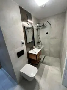 Nowoczesna łazienka wyposażona w kabinę prysznicową ze szklaną przegrodą, toaletę montowaną na ścianie, drewnianą toaletkę z umywalką oraz elegancką czarną armaturę uzupełnioną dużymi szarymi płytkami.