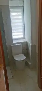 Nowoczesna łazienka z marmurowymi płytkami, wyposażona w czystą białą toaletę obok szklanej przegrody prysznicowej, z dobrze oświetlonym oknem zapewniającym naturalne światło.
