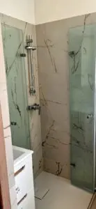Nowoczesny narożnik łazienkowy z przeszklonym prysznicem, marmurowymi panelami ściennymi i eleganckim systemem prysznicowym.